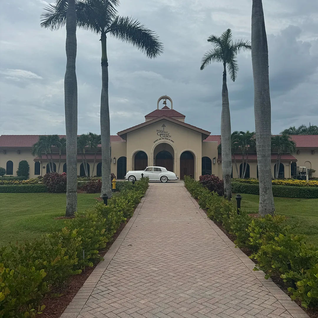 Vintage white Rolls Royce Silver Cloud II parked at St. Leo the Great Catholic Church beyond a palm-lined walkway in Bonita Springs, Florida