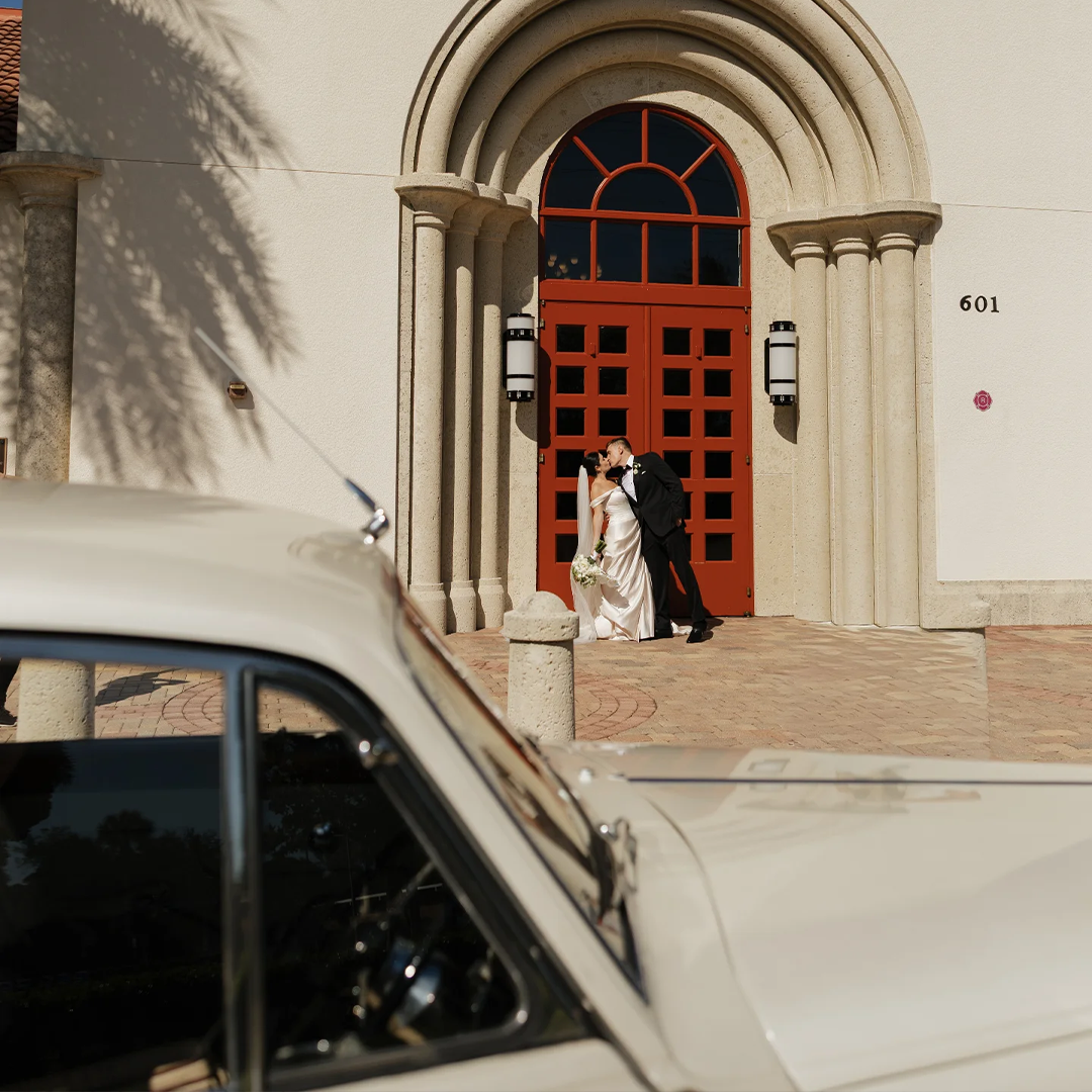 Saint William Catholic Church Naples FL Wedding Rolls Royce Silver Cloud II Bride and groom kissing beneath the red arched doors of Saint William Catholic Church with the vintage cream white Rolls Royce Silver Cloud II in Naples Florida