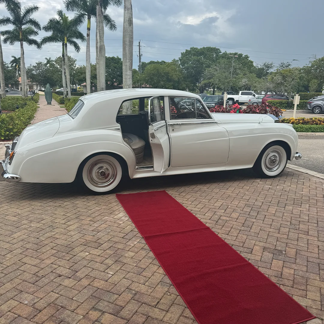 Morelli Wedding Red Carpet Rolls-Royce Vineyards Naples FL Vintage cream white 1960 Rolls-Royce Silver Cloud II parked on a paver drive with a long red carpet leading to the open rear door at Vineyards Country Club in Naples Florida