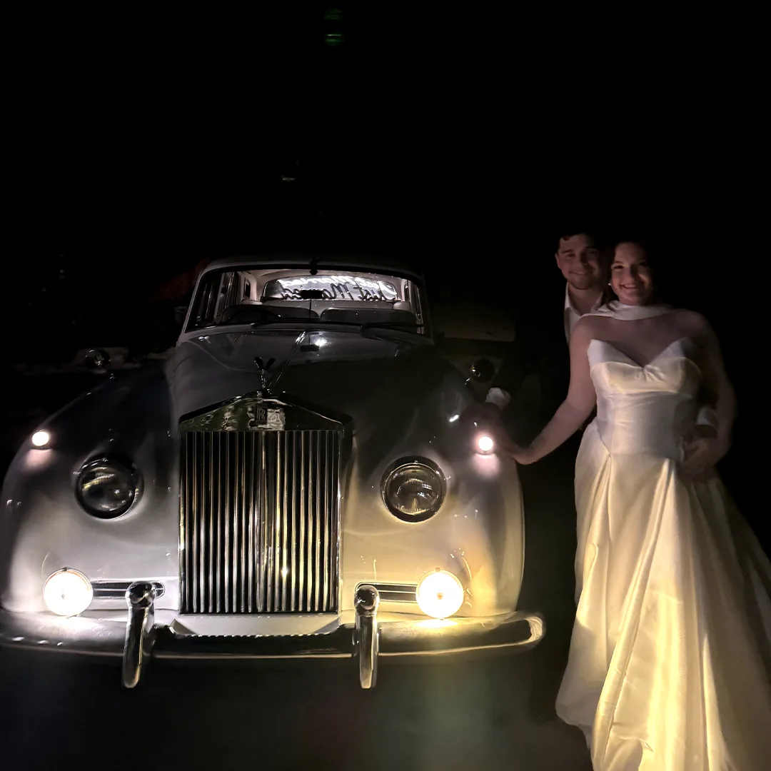 Bride and groom standing beside the glowing headlights of a vintage white Rolls Royce Silver Cloud II at night after their Palmira Golf Club wedding in Bonita Springs, Florida