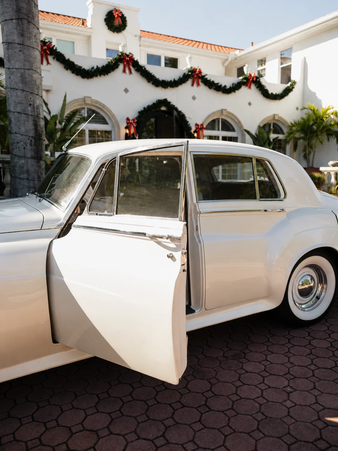 Shangri-La Springs wedding venue — open rear door of the cream-white 1960 Rolls-Royce Silver Cloud II in front of the holiday-decorated façade in Bonita Springs, Florida