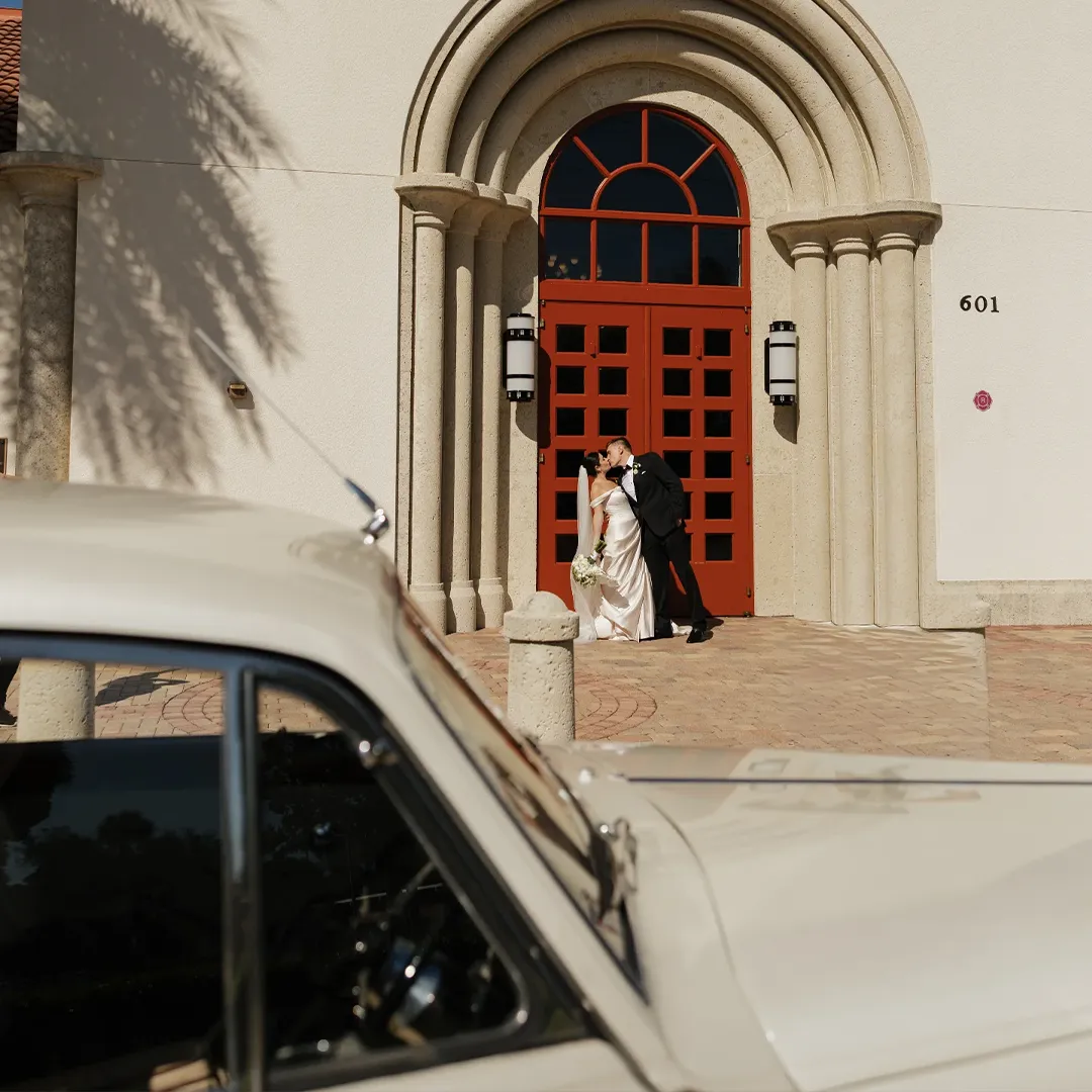 Saint William Catholic Church Naples FL Wedding Rolls Royce Silver Cloud II Bride and groom kissing beneath the red arched doors of Saint William Catholic Church with the vintage cream white Rolls Royce Silver Cloud II in Naples Florida