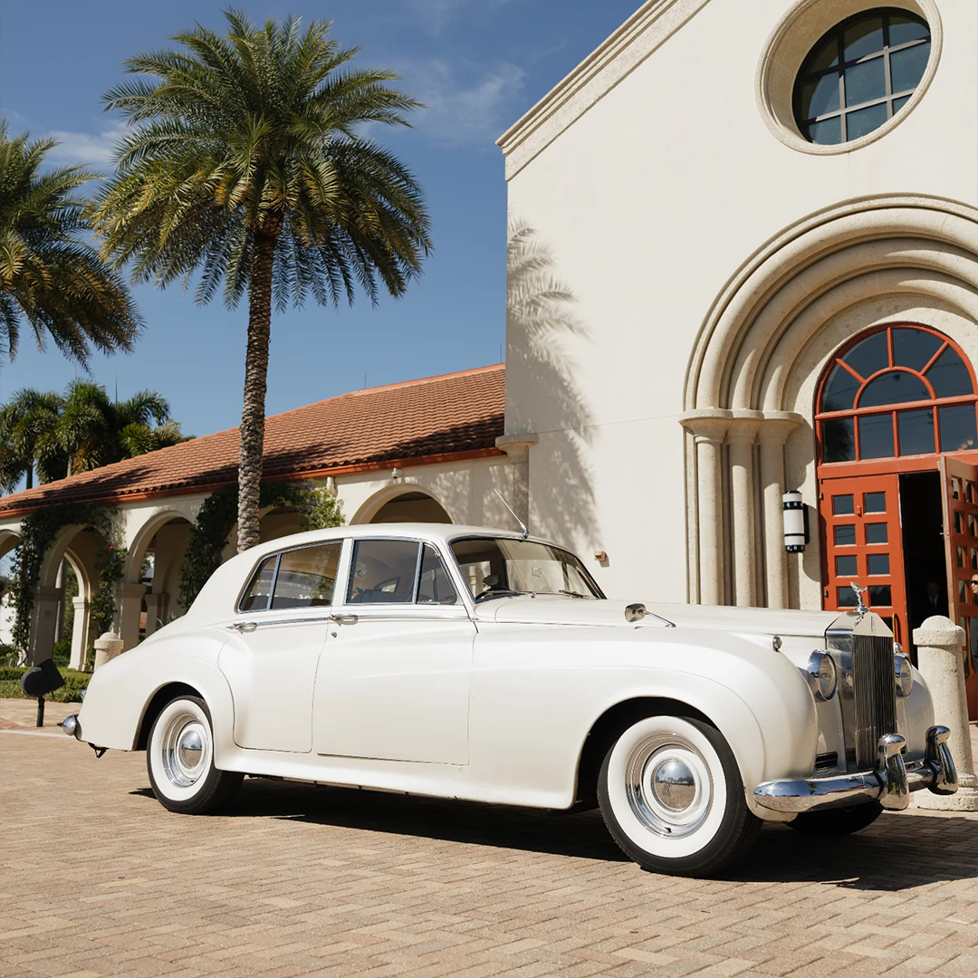 Front quarter view of a vintage cream white Rolls Royce Silver Cloud II framed by palm trees at Saint William Catholic Church in Naples, Florida