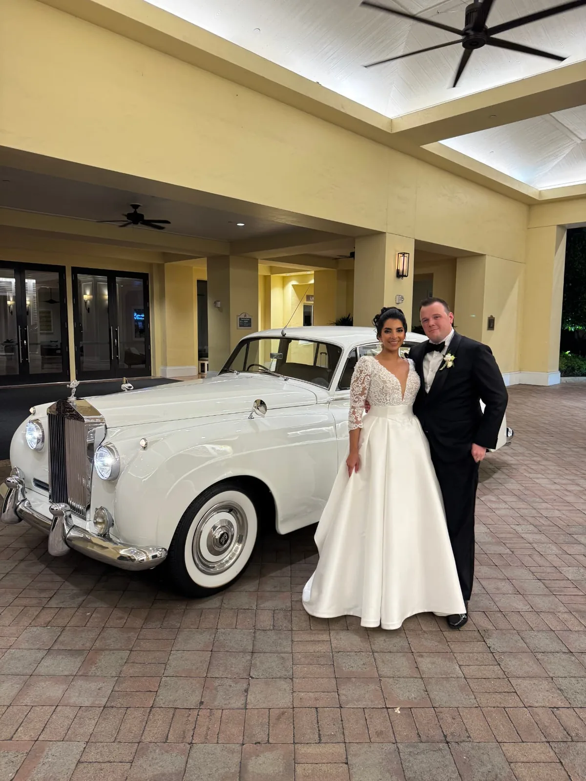 Bride and groom in a lace gown and black tuxedo standing beside a cream white vintage Rolls-Royce Silver Cloud II under the porte cochere at The Club at Grandezza in Estero Florida