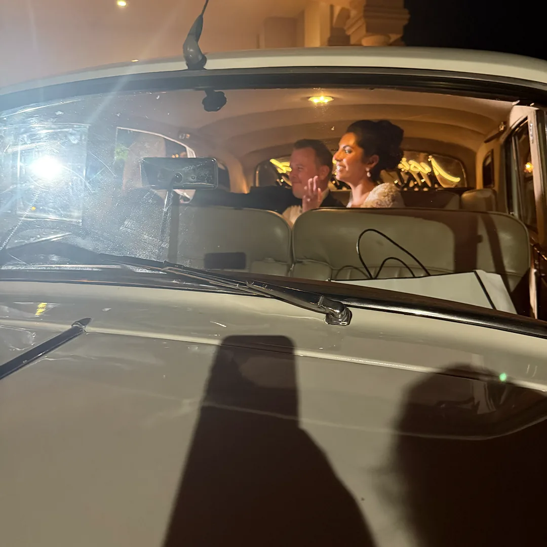 Bride in a beaded gown and groom in a black tuxedo waving from the back seat of the vintage cream white Rolls Royce Silver Cloud II seen through the windshield at night with a glowing Just Married sign behind them, after the Southerland wedding at The Club at Grandezza in Estero, Florida
