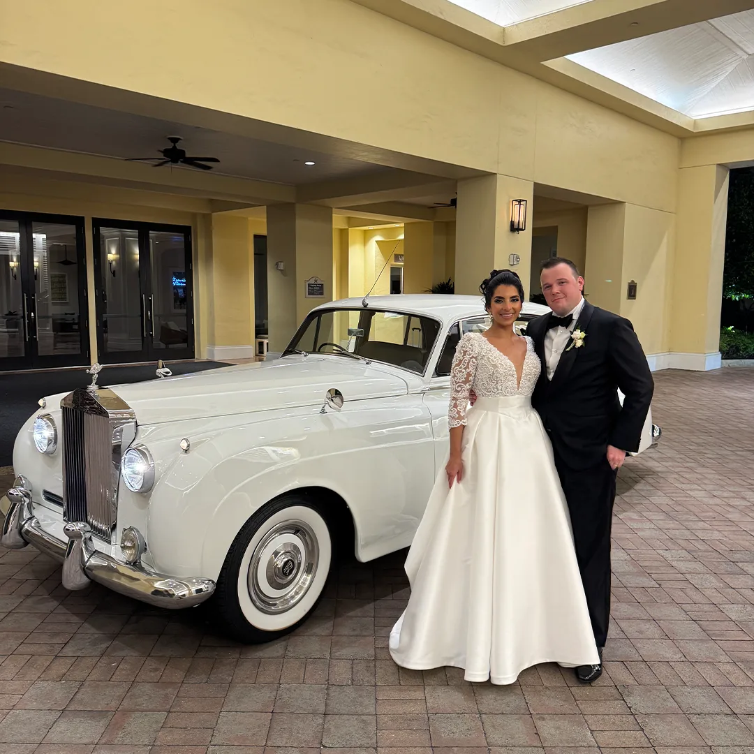 Bride in a lace gown and groom in a black tuxedo standing beside a vintage white Rolls Royce Silver Cloud II beneath the porte cochere at The Club at Grandezza in Estero, Florida
