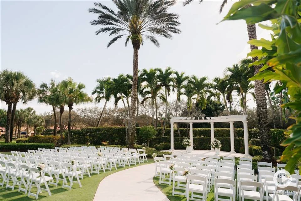 The Club at Grandézza wedding venue — outdoor garden ceremony with white chairs, palm trees, and a white pergola in Estero, Florida