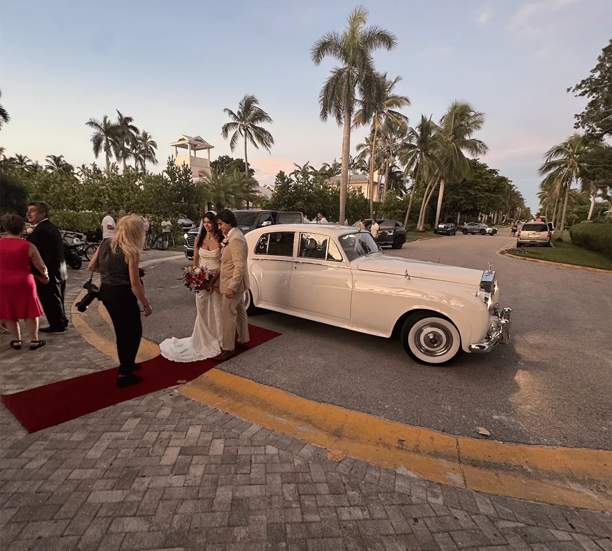 Bride and groom standing on a red carpet beside a vintage white Rolls Royce Silver Cloud II at their beach wedding on 8th Ave South in Naples, Florida