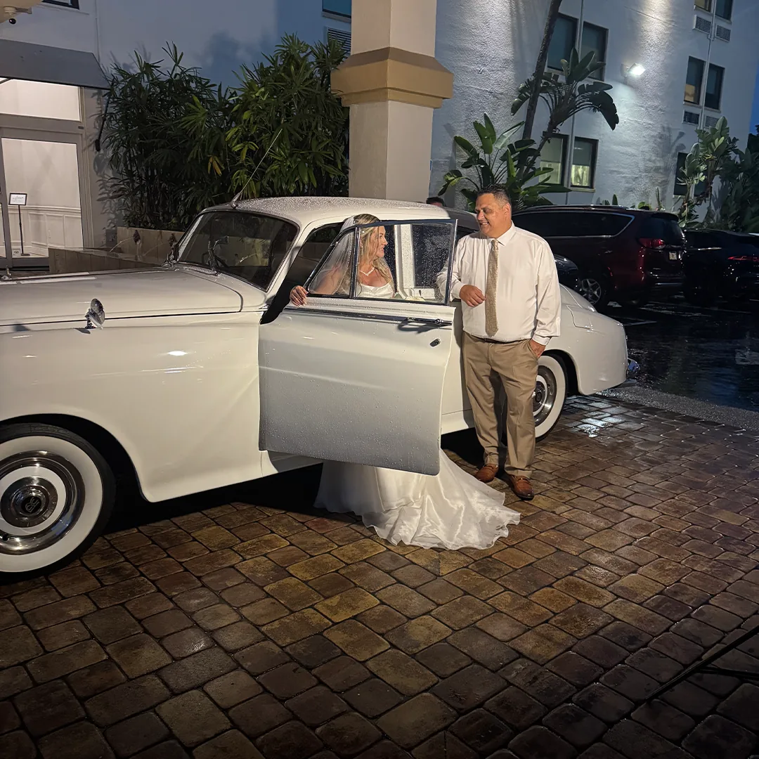 Bride in a strapless gown stepping from the open door of a vintage white Rolls Royce Silver Cloud II on a wet brick driveway at the Bayfront Inn in Naples, Florida at night while a groomsman in a tan suit stands beside the car