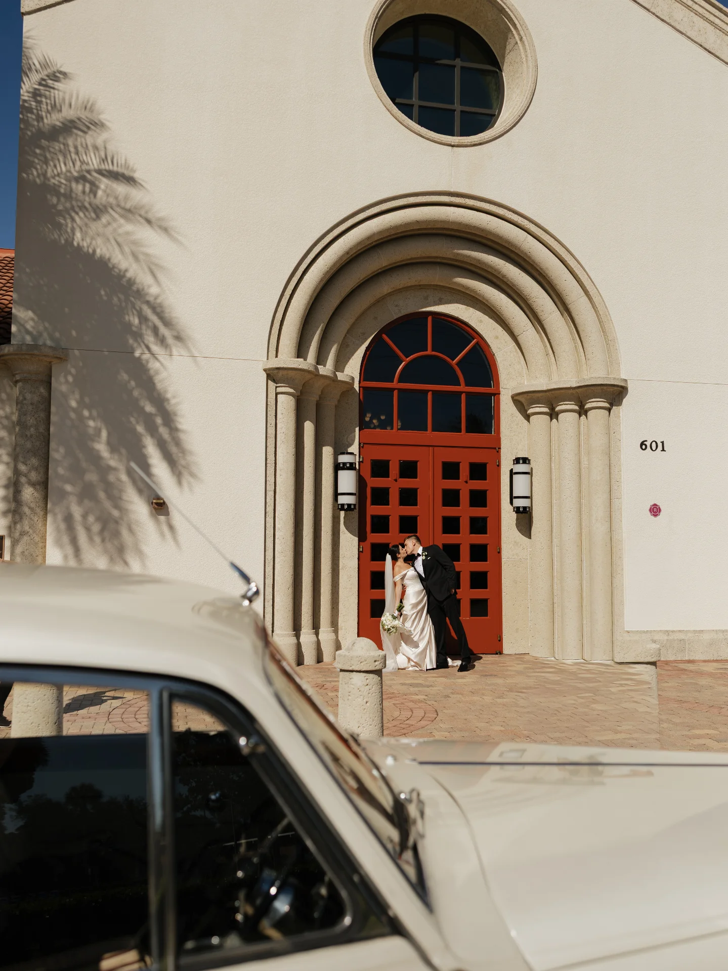 Saint William Catholic Church wedding — bride and groom kissing beneath the red doors with the vintage Rolls-Royce at the Naples venue