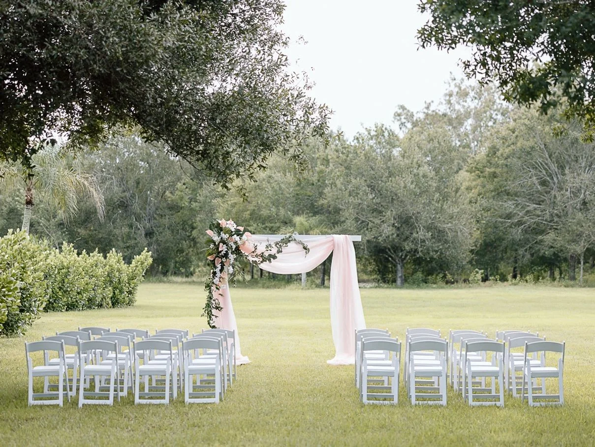 Rock Manor wedding venue — outdoor ceremony lawn with white chairs and a floral draped arch beneath mature trees in Ft. Myers, Florida
