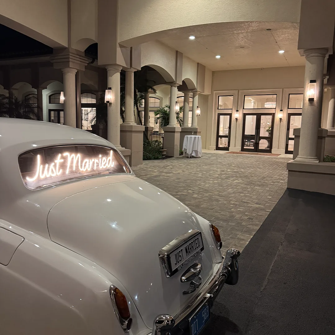 The Club at Grandézza Wedding — Estero, FL The Club at Grandézza wedding — cream-white 1960 Rolls-Royce Silver Cloud II with a glowing 'just married' script beneath the lit porte-cochère at night in Estero, Florida